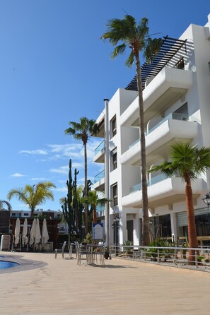 A modern building with multiple balconies, palm trees, and a pool area with umbrellas.