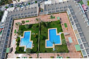 An aerial view of a hotel with two swimming pools, surrounded by palm trees and a parking area.