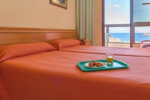 A hotel room with two beds, a breakfast tray with pastries and juice, and a view of the sea through large windows.