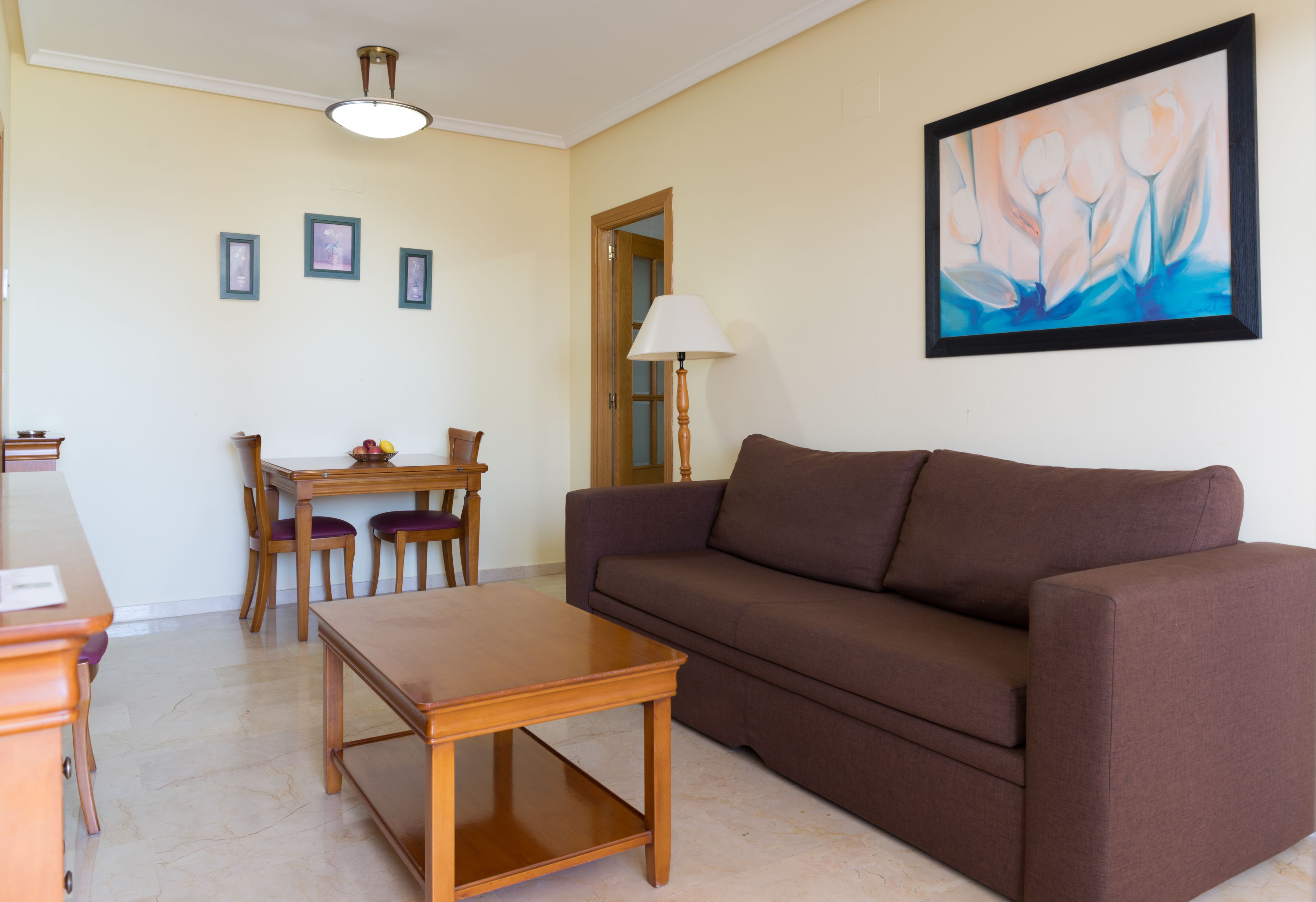 A living room with a brown sofa, wooden coffee table, and a dining table with chairs. There is a painting on the wall and a lamp near the dining area.