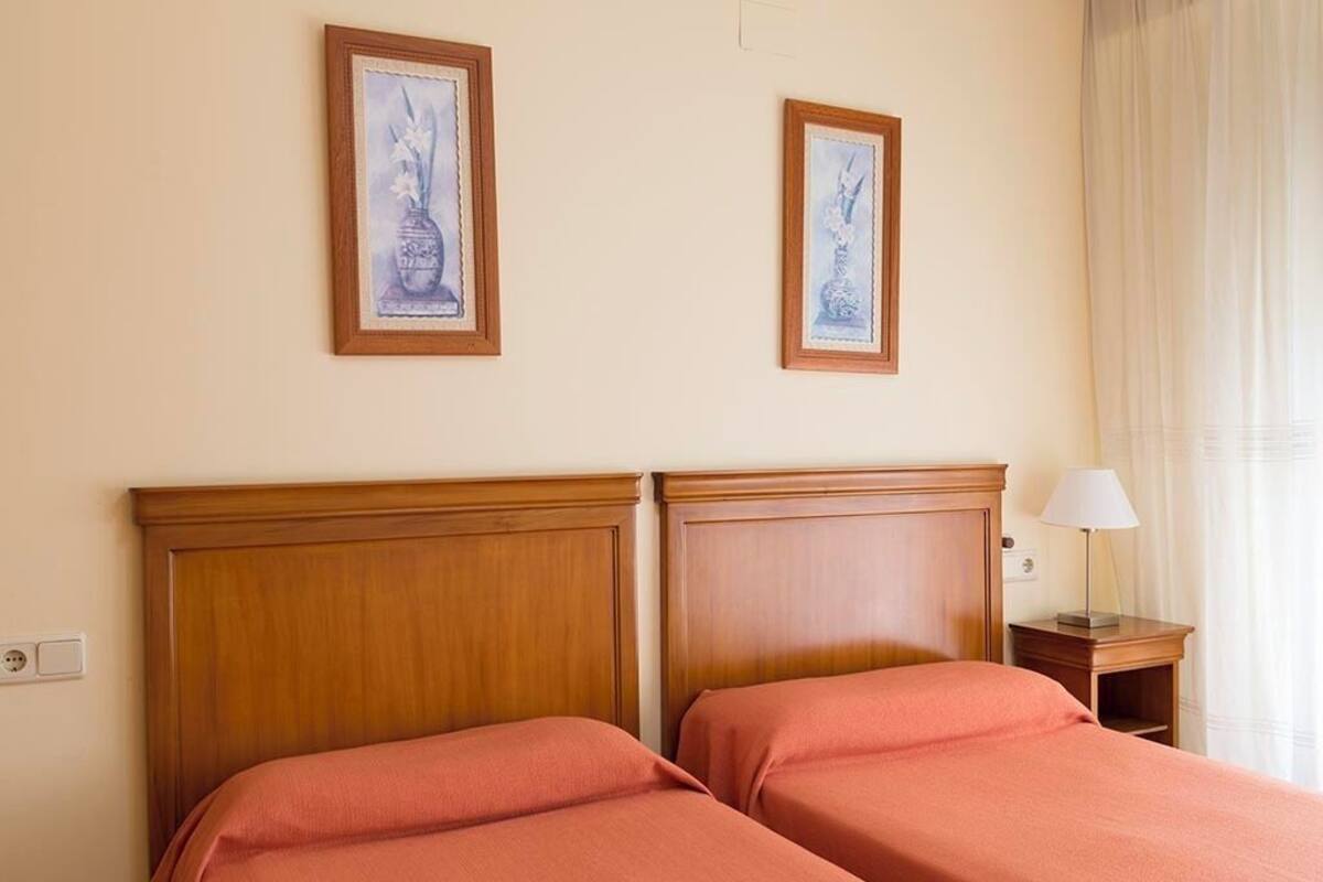 Ein Hotelzimmer mit zwei hölzernen Betten, einem Nachttisch mit Lampe und zwei gerahmten Kunstwerken an der Wand.