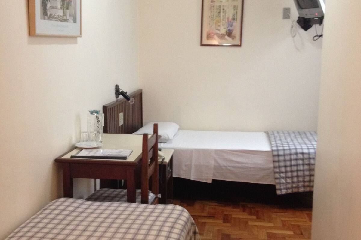 A hotel room with two beds, a desk, a chair, a television, and two framed pictures on the wall.