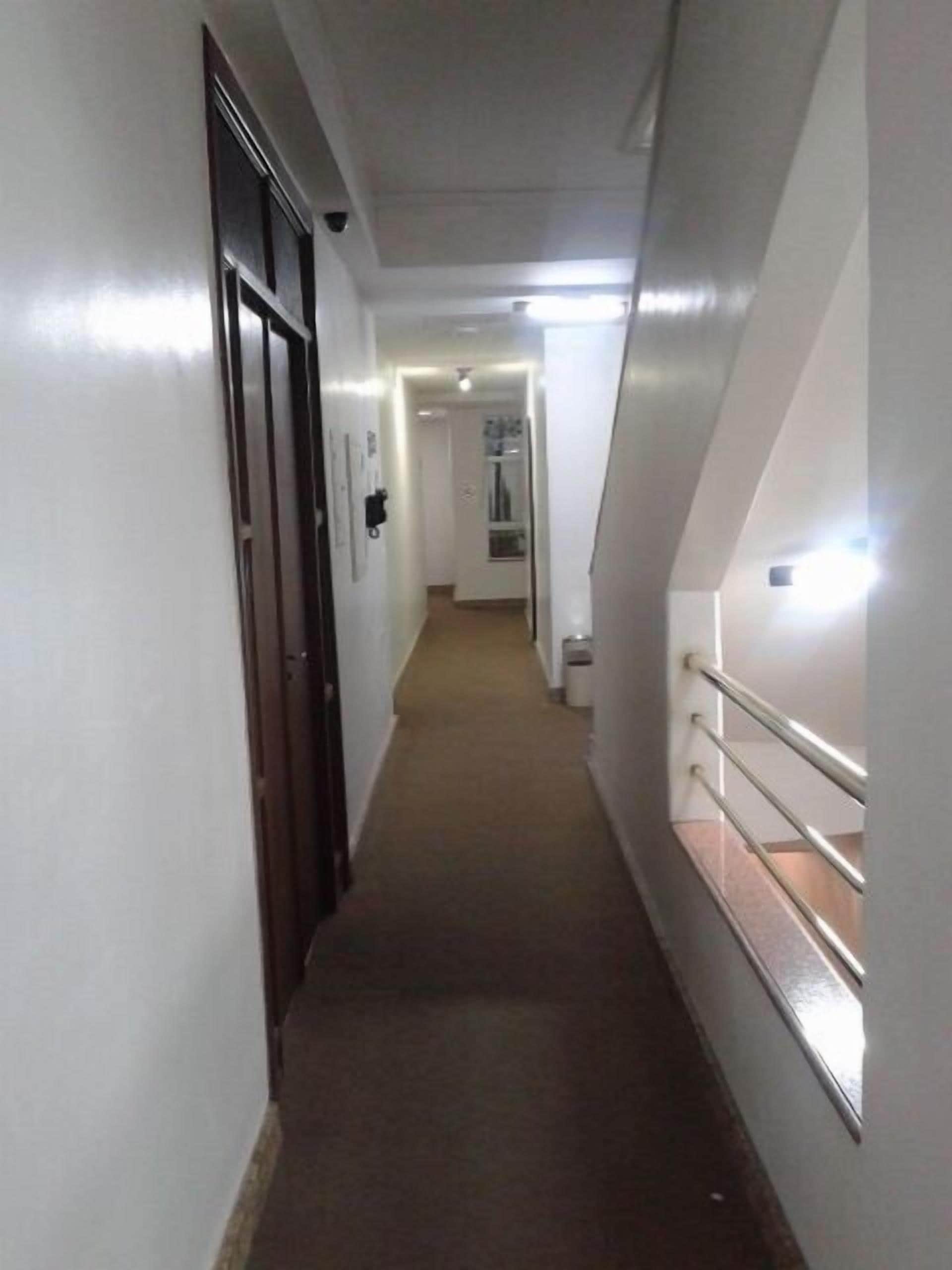 A narrow, well-lit hallway with a carpeted floor, a wooden door on the left, and a metal railing on the right.