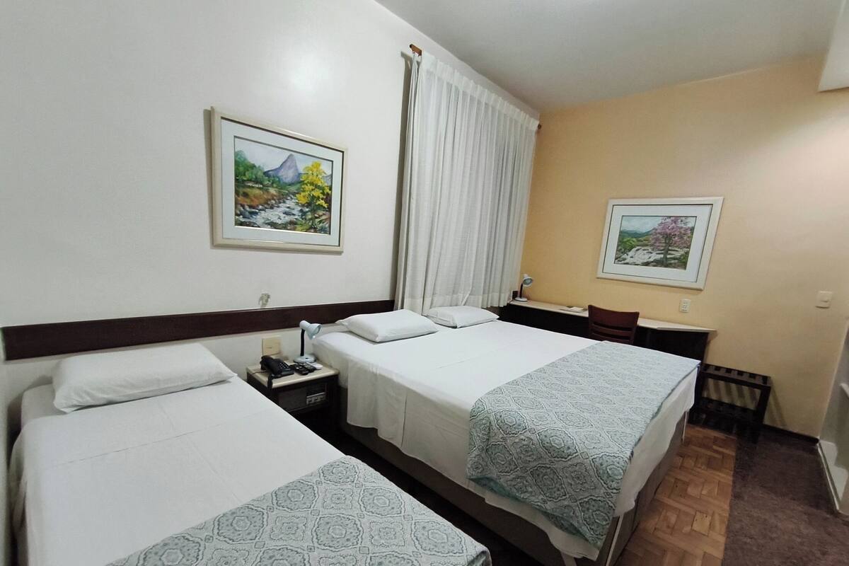 A hotel room with two beds, a desk, a chair, a nightstand with a lamp, and two framed pictures on the walls.