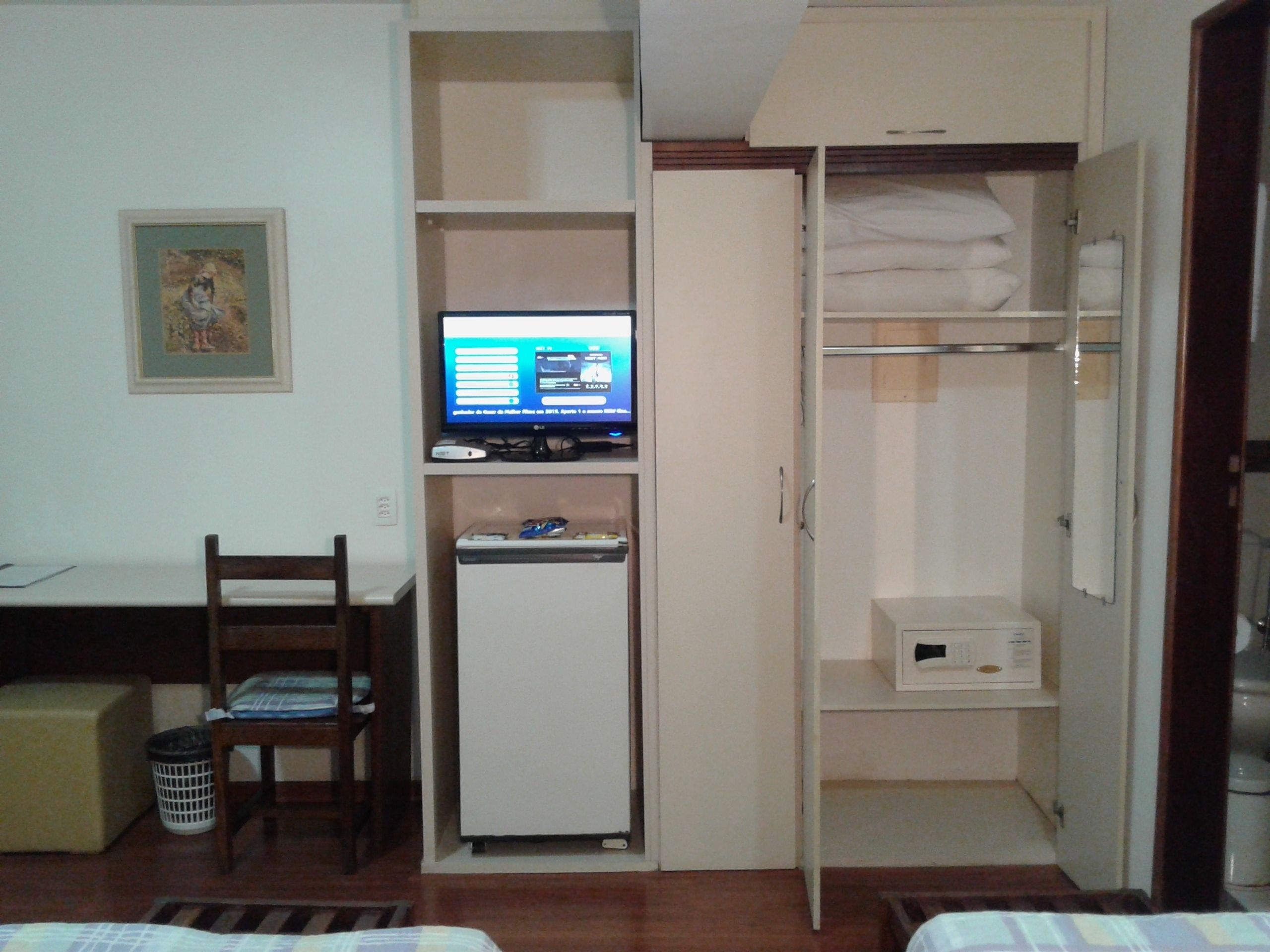 Habitación compacta con cama, escritorio con computadora, refrigerador pequeño y armario con espacio para colgar.