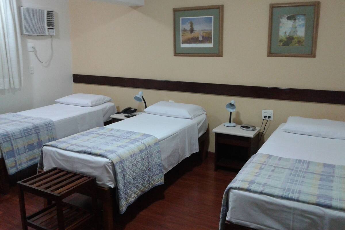 A hotel room with two single beds, a wooden bench, a bedside table with a lamp, and two framed pictures on the wall.