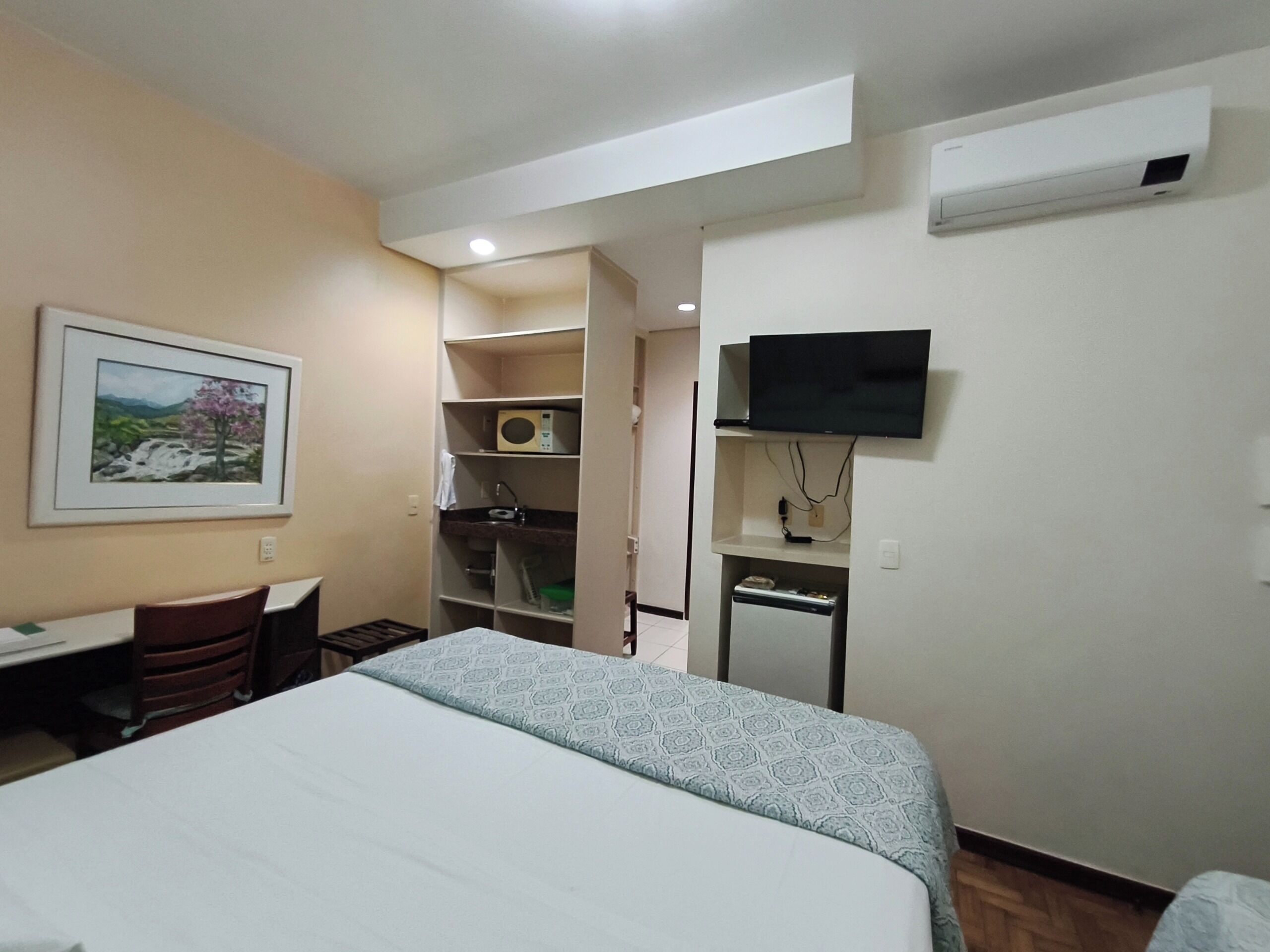 A hotel room with a bed, a desk, a chair, a TV, and a kitchenette.