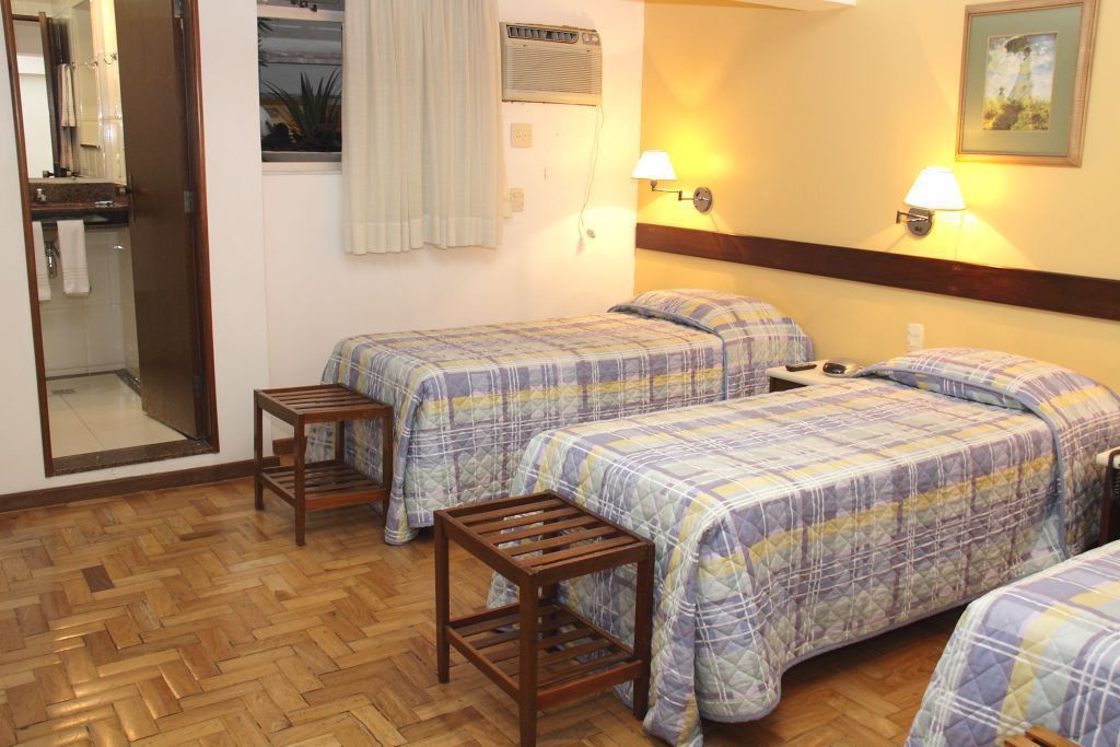 Habitación de hotel con dos camas, muebles de madera, una mesita, un espejo, una ventana con cortinas y un calefactor en la pared.
