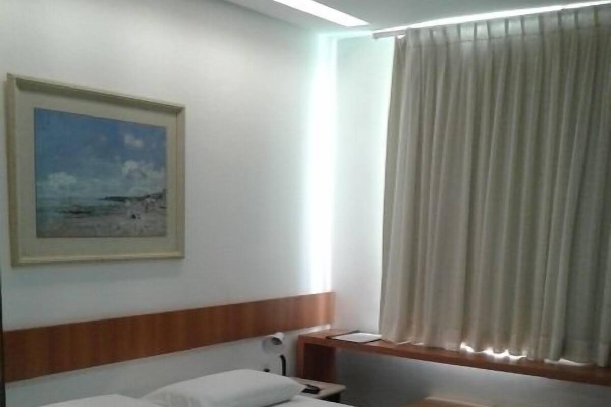 A hotel room with a bed, a nightstand, a painting, a window with curtains, and a wooden headboard.