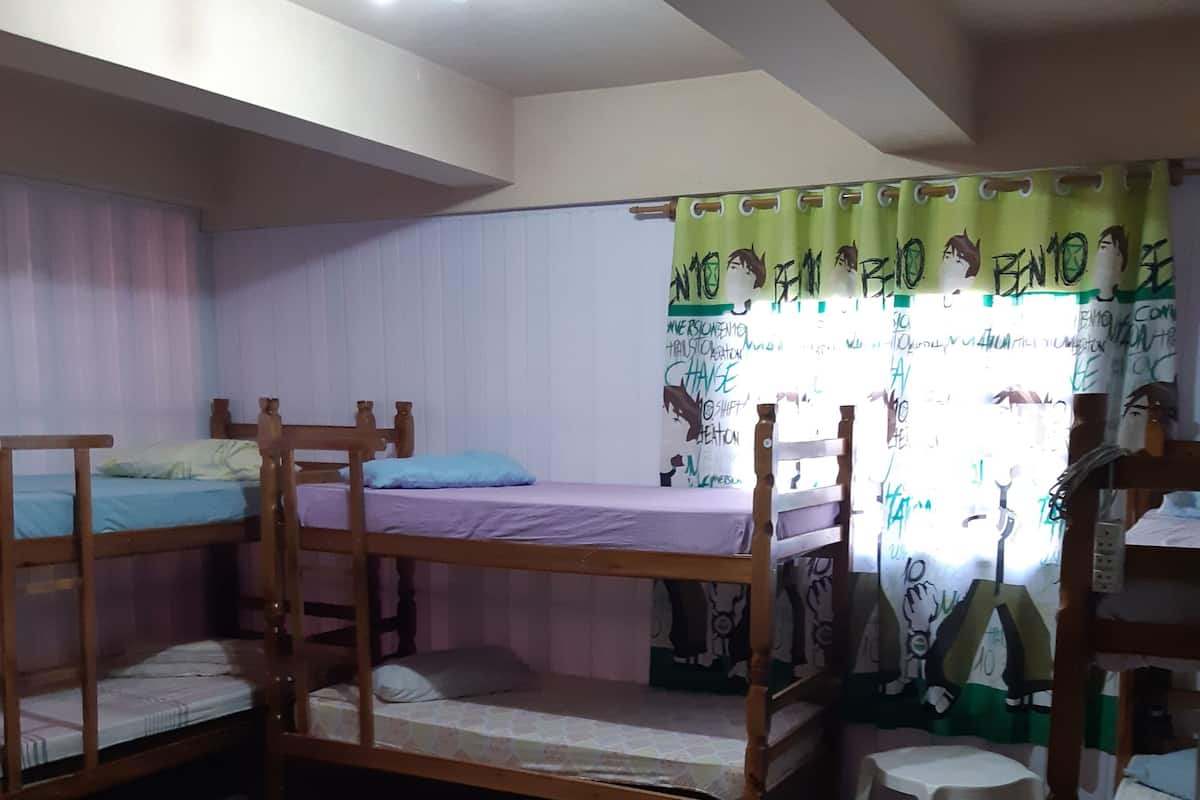 Une chambre de dortoir avec des lits superposés, un petit tabouret et une fenêtre avec un rideau.