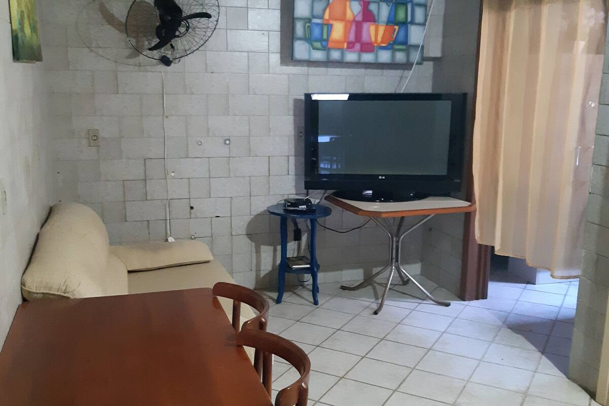 Un espace de vie compact doté d’un sol carrelé, d’une table à manger en bois, d’un canapé, d’un téléviseur posé sur un meuble et d’un ventilateur fixé au mur.