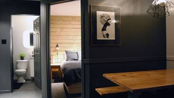 A modern hotel room with a wooden floor, a bed with a grey and black bedspread, a bedside table, a framed picture on the wall, and a wooden bench at the table.