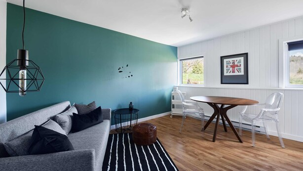 A modern living room with a teal accent wall, a grey sofa with black cushions, a round wooden table with chairs, and a black and white striped rug.