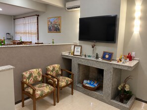 A living room with a flat-screen TV, two floral-patterned chairs, a fireplace with magazines, and a framed certificate on the mantel. - Pousada Canasvieiras (Florianópolis)