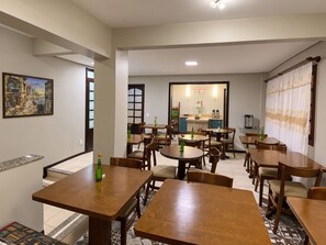 A dining area with multiple wooden tables and chairs, a kitchen in the background, and a painting on the wall. - Pousada Canasvieiras (Florianópolis)