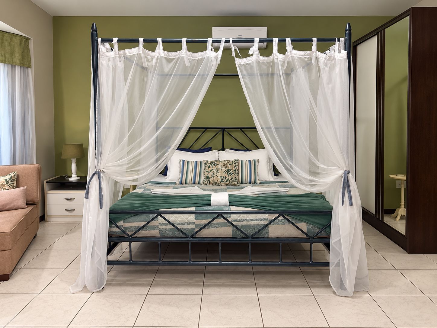 Un lit avec un cadre en métal, un ciel de lit en tissu blanc et une literie verte. Il y a une table de chevet avec une lampe, ainsi qu’une petite table avec une autre lampe.