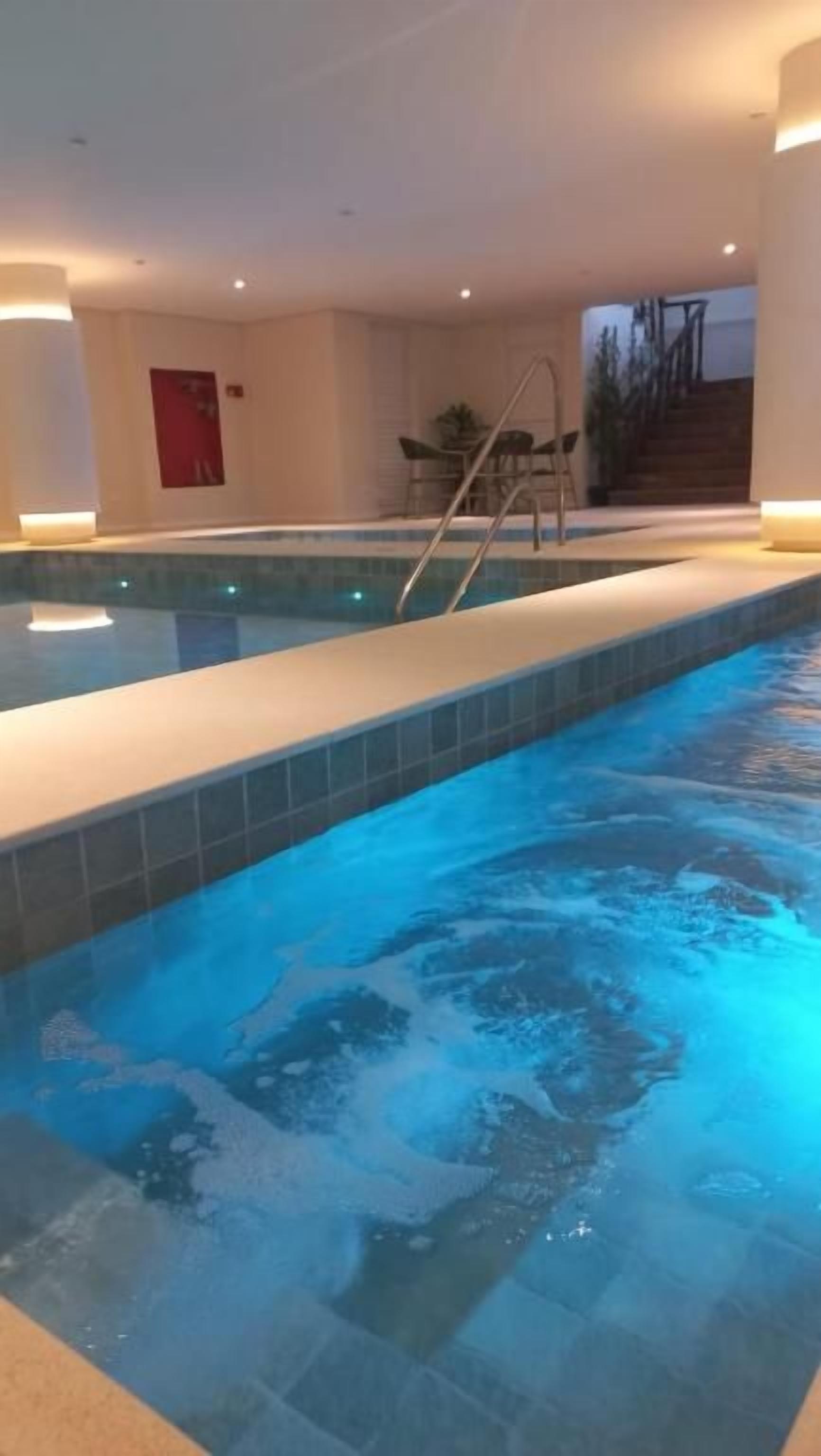 An indoor swimming pool with a ladder, tiled walls, and a staircase in the background.