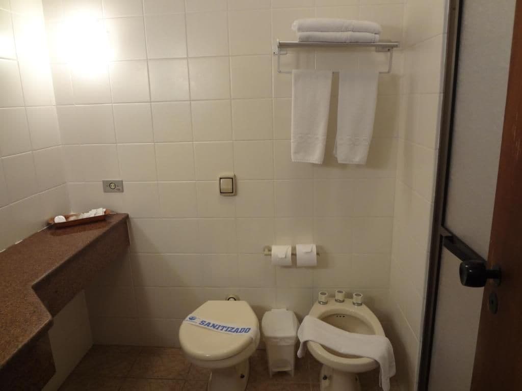 Un baño pequeño con dos inodoros, uno de ellos con la etiqueta 'SANITIZED'. Hay un perchero con toallas sobre el inodoro, y un pequeño cubo de basura es visible.