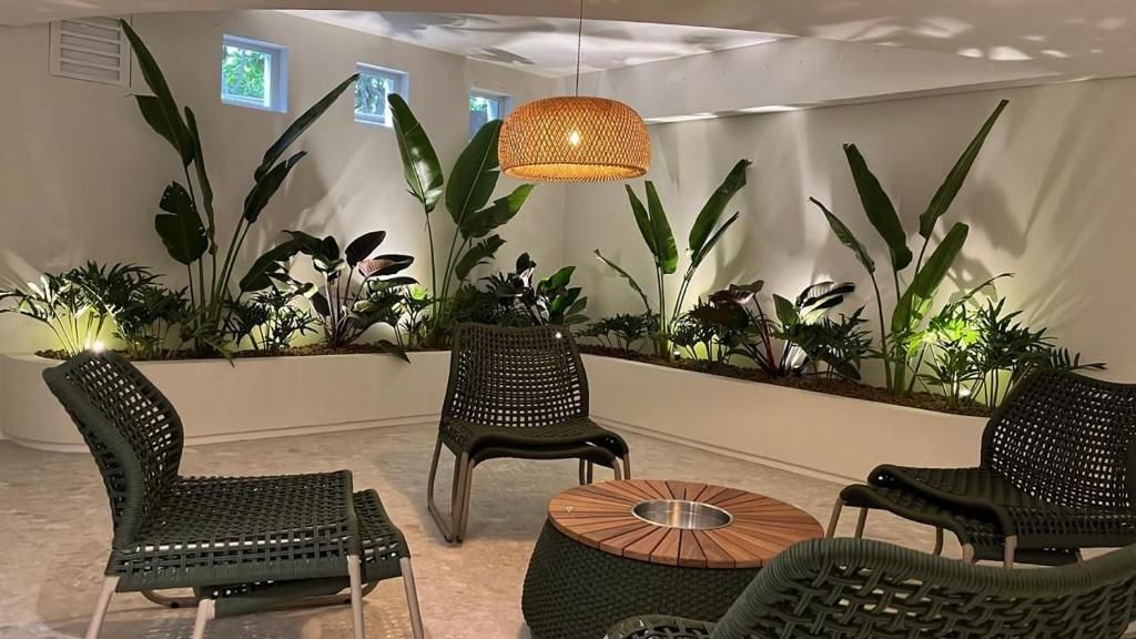 A room with a seating area featuring wicker chairs and a round table, surrounded by large potted plants.