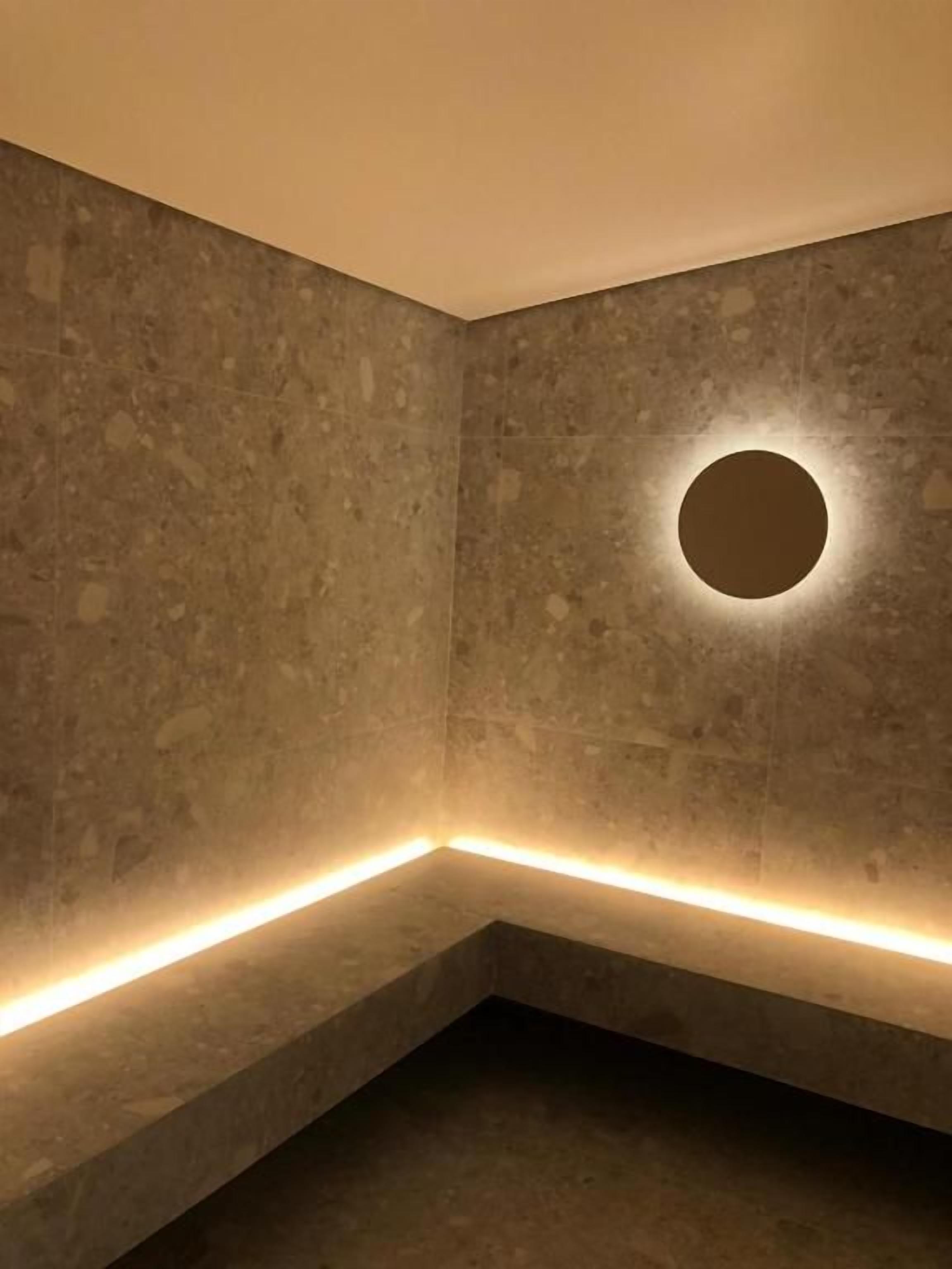 Canto de um quarto com luminária circular na parede e fita de luz ao longo do rodapé.