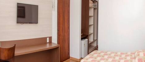 Quarto com escrivaninha de madeira, televisão na parede, guarda-roupa e cama com colcha.