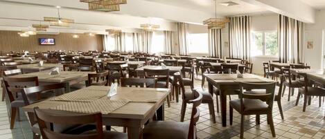 Un vaste espace dédié à la salle à manger, avec plusieurs tables et chaises, un écran de télévision et de grandes fenêtres laissant entrer la lumière naturelle.