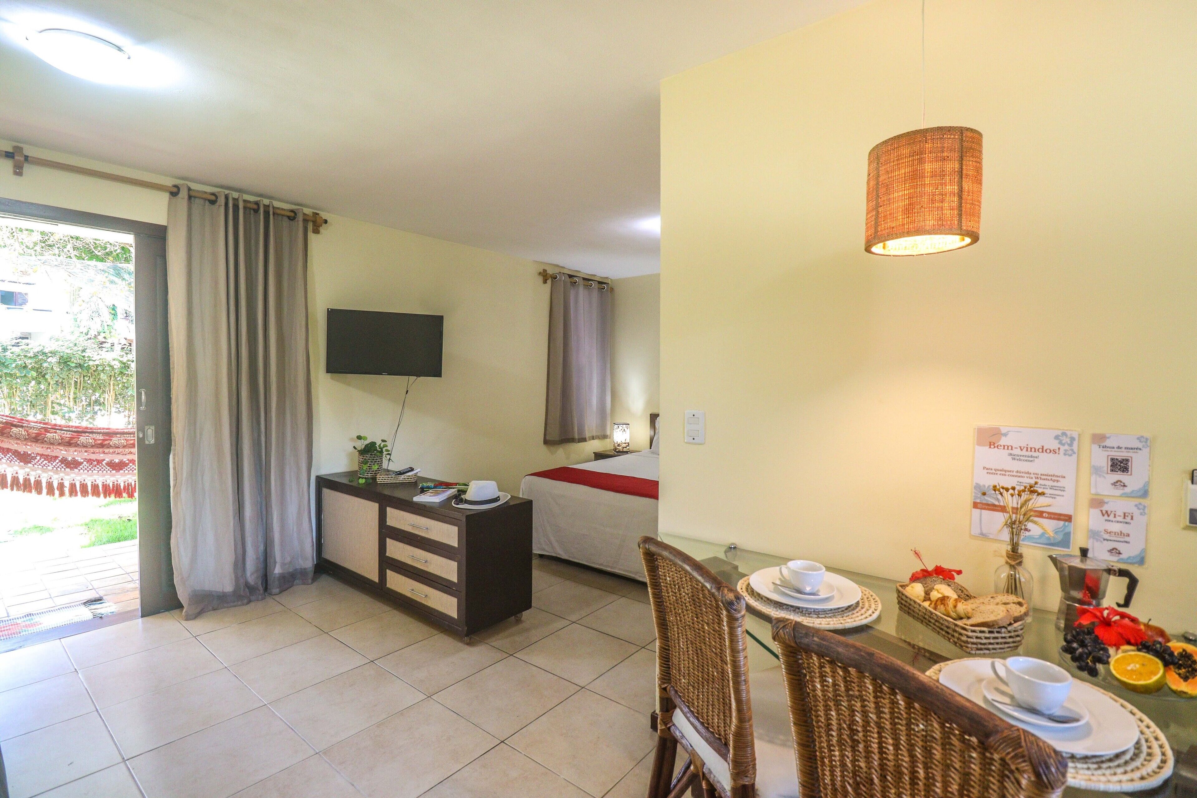 Quarto de hotel com cama, escrivaninha, televisão, área de refeições com mesa e cadeiras, e vista para o exterior.