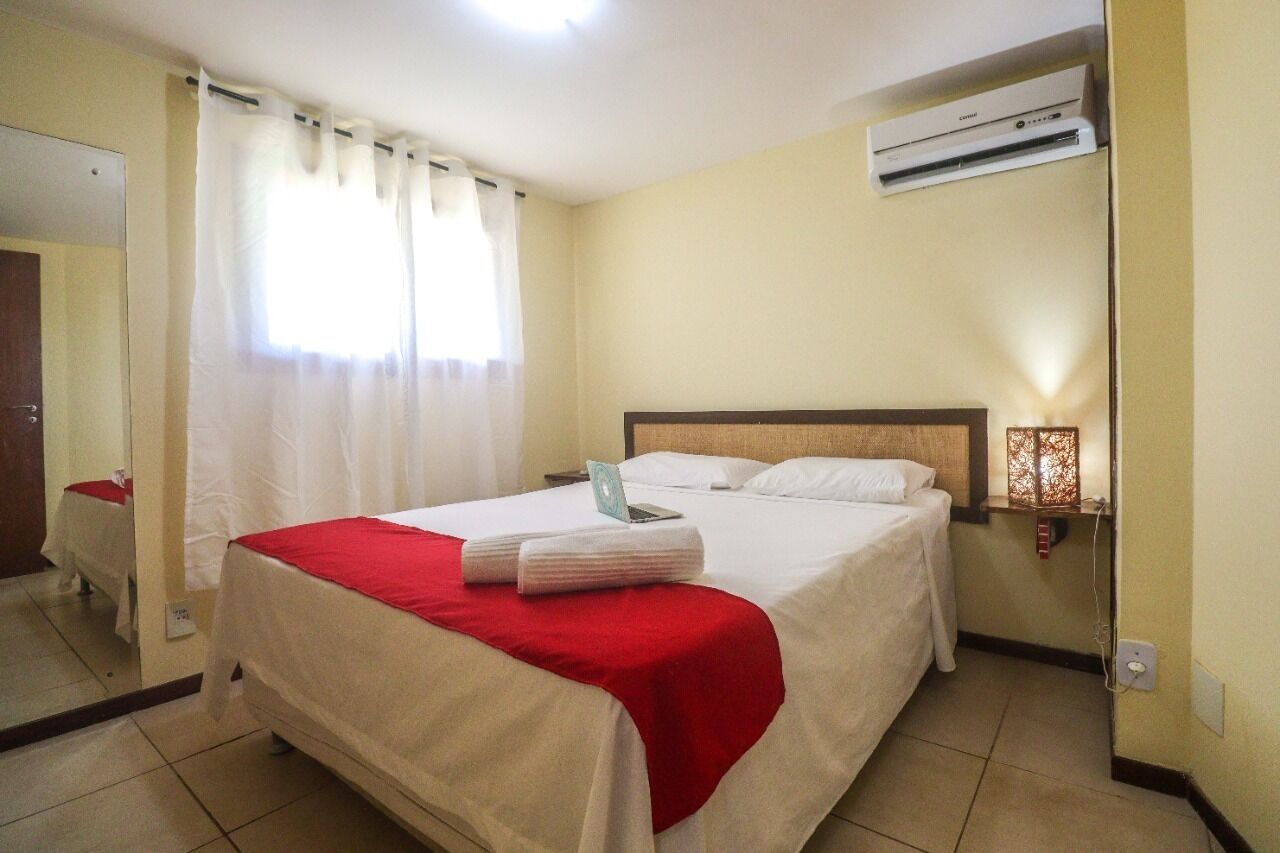 A hotel room with a bed, a laptop, a red and white bedspread, an air conditioner, and a nightstand with a lamp.