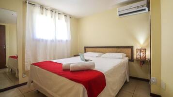 A hotel room with a bed, a laptop, a red and white bedspread, an air conditioner, and a nightstand with a lamp.