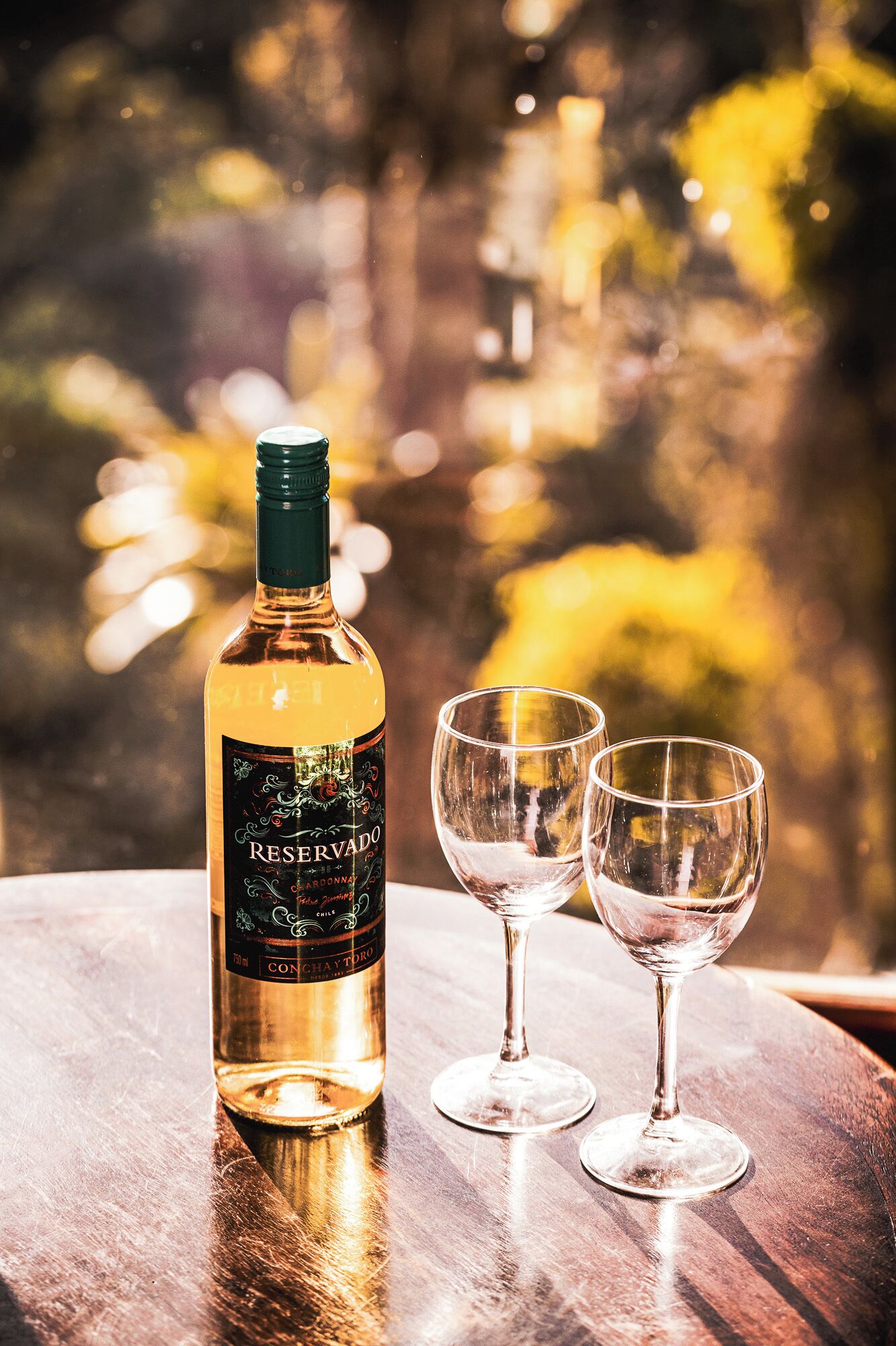 A bottle of Reserva wine and two empty wine glasses on a wooden table with a blurred outdoor background.