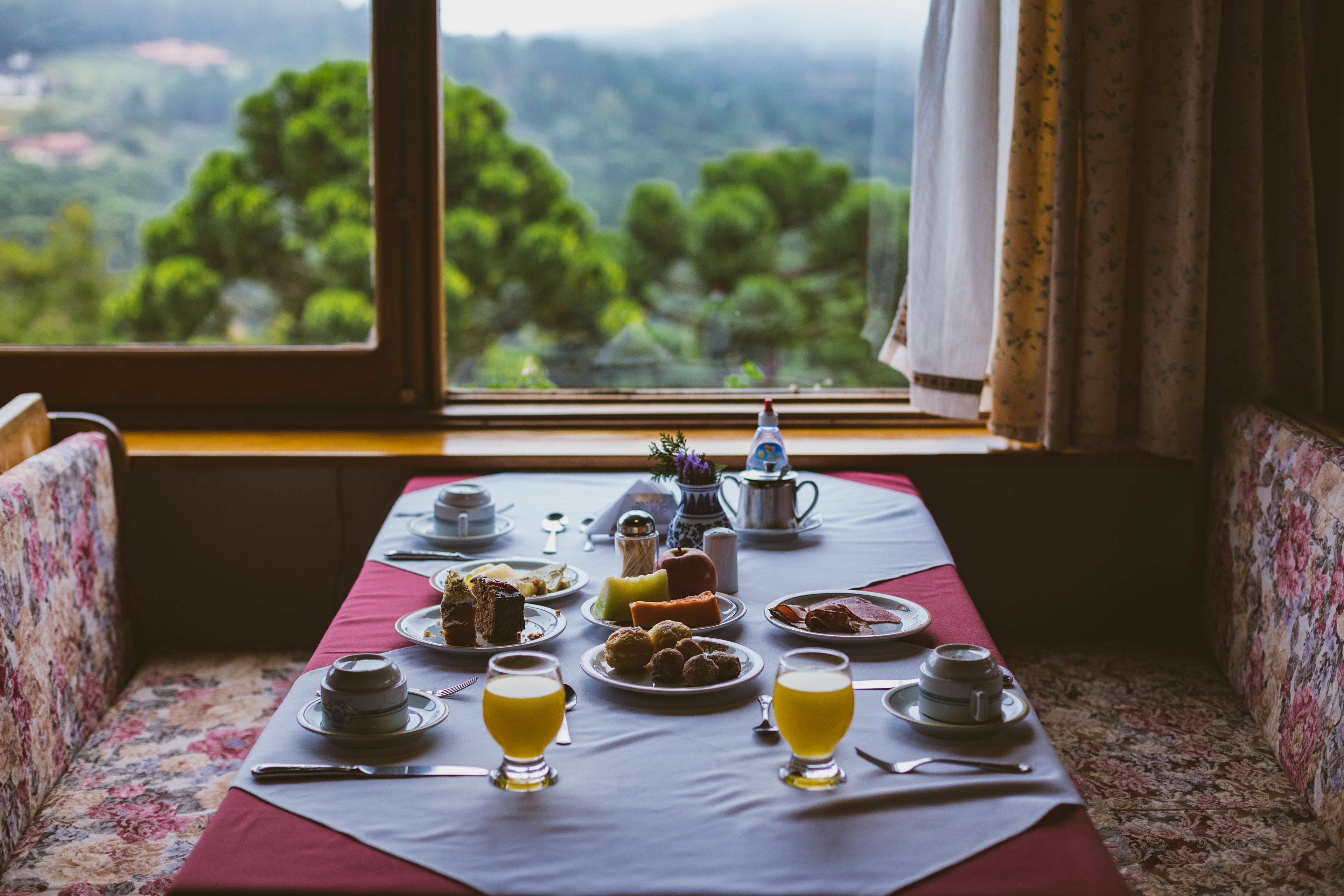 A breakfast table set with various dishes and drinks, a window with a view of green trees, and floral-patterned upholstery.