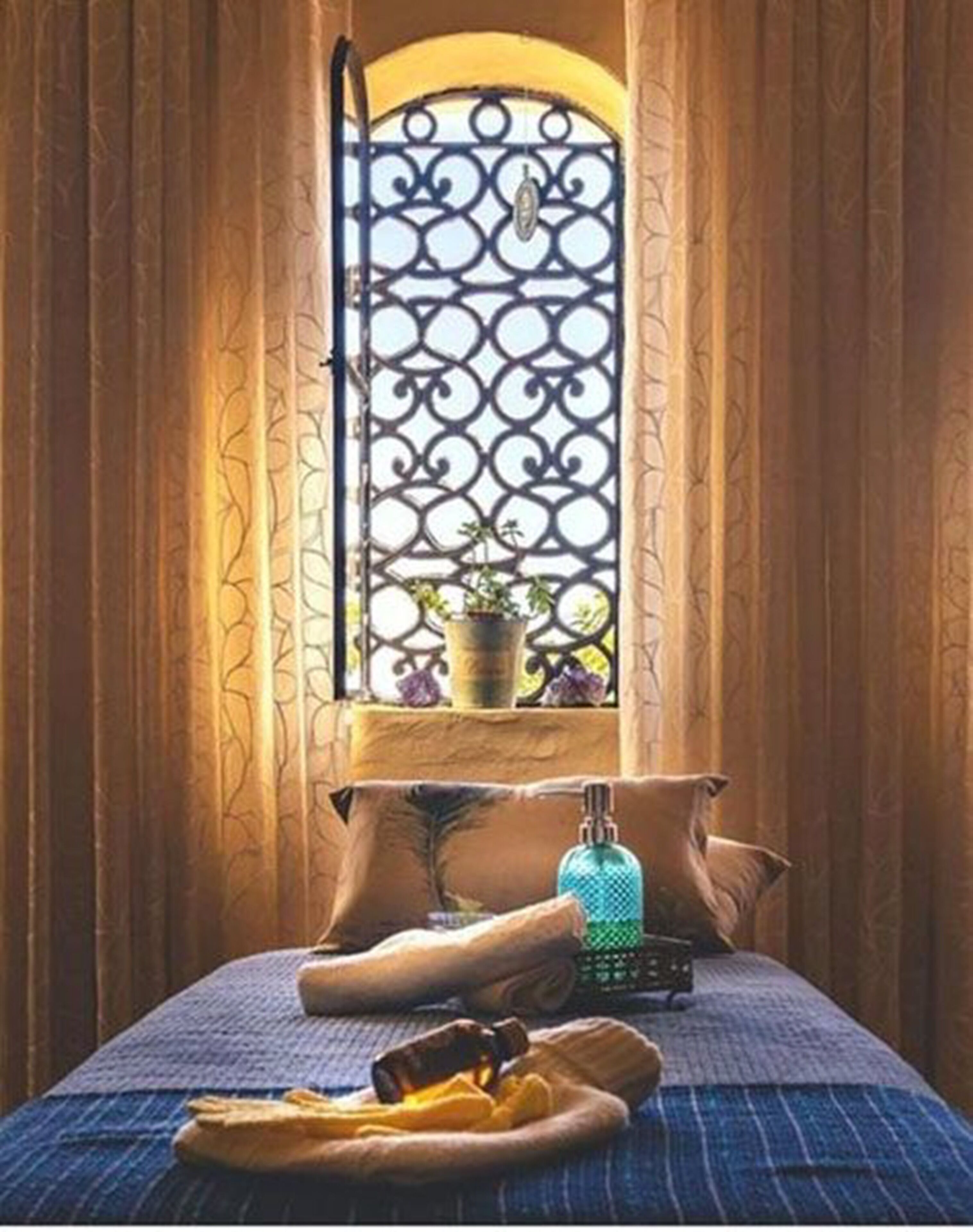 Una stanza con un letto, una finestra con inferriate decorative e uno scaffale con una pianta e una bottiglia.