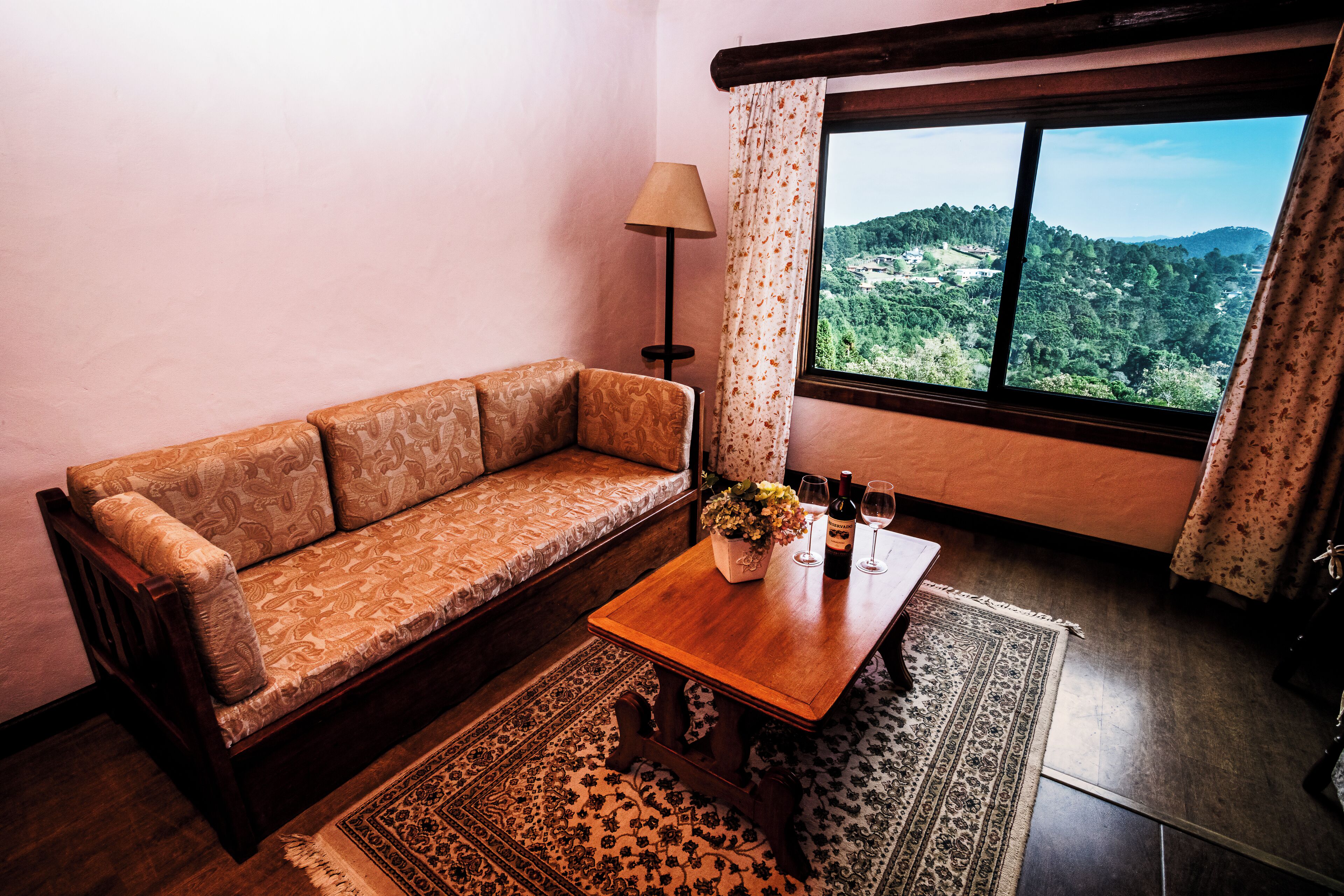 A cozy living room with a sofa, a wooden coffee table with flowers and wine glasses, a lamp, and a view of a forested hillside through a window.