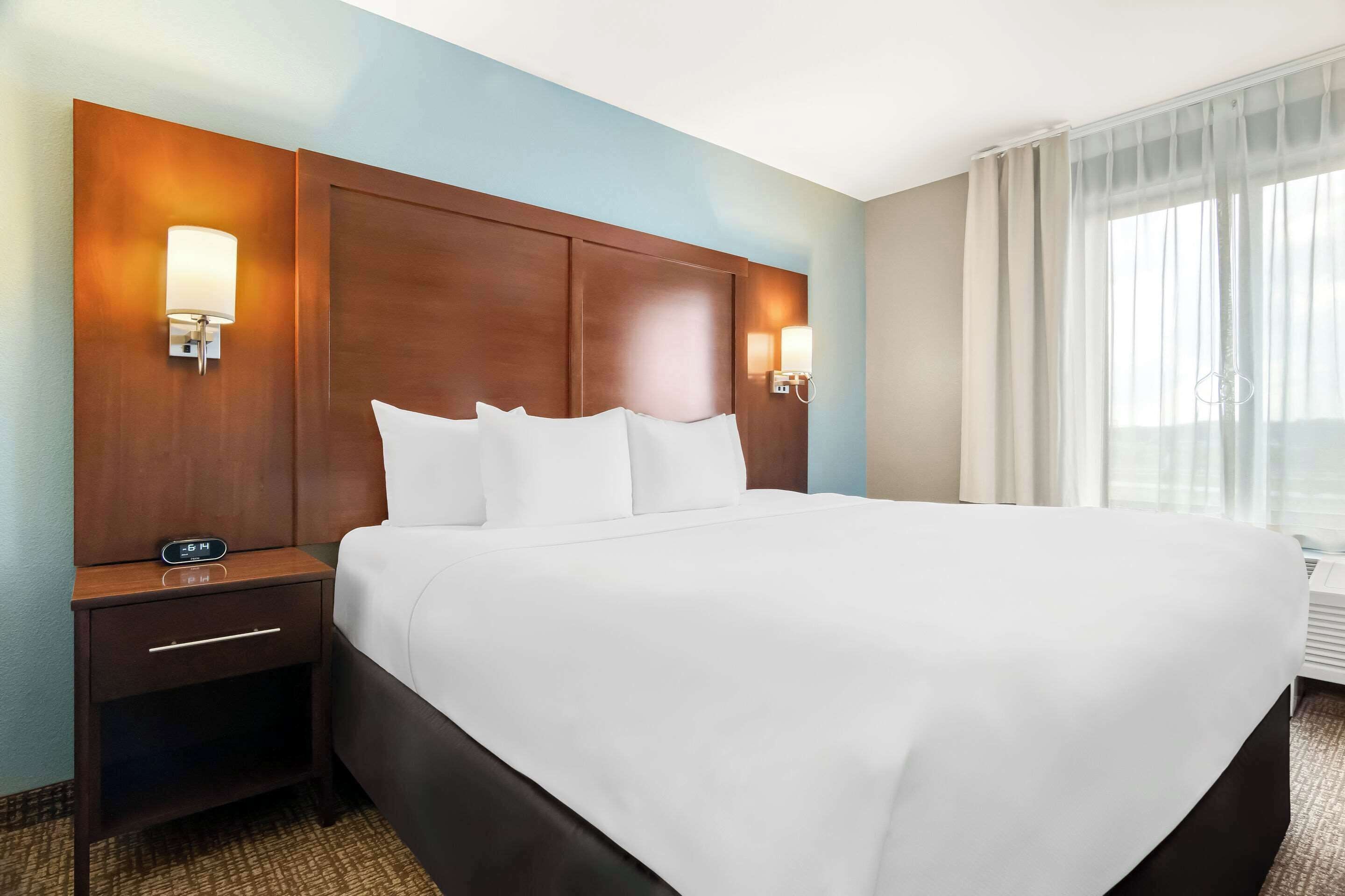 A hotel room with a large bed, wooden headboard, two bedside lamps, a nightstand with a clock, and a window with sheer curtains.