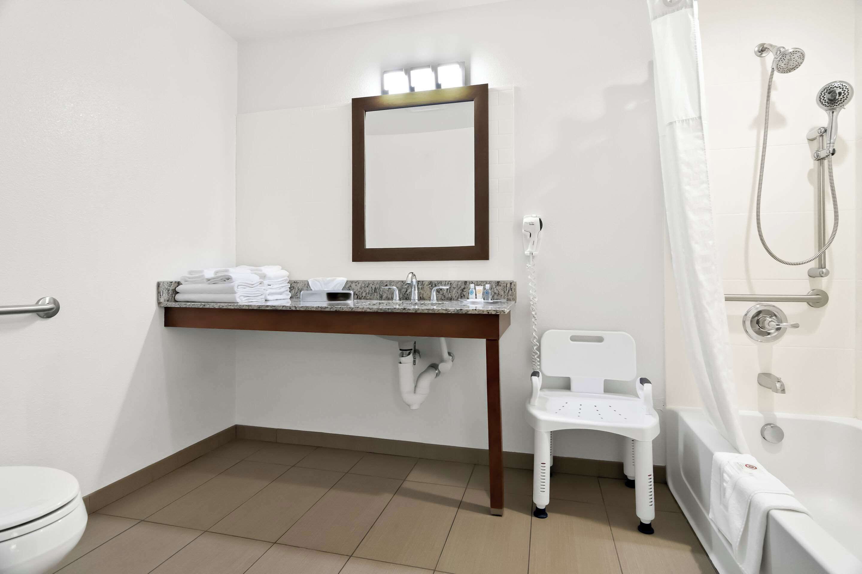 A bathroom with a toilet, sink, mirror, shower, and a white chair.