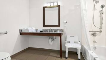 A bathroom with a toilet, sink, mirror, shower, and a white chair.