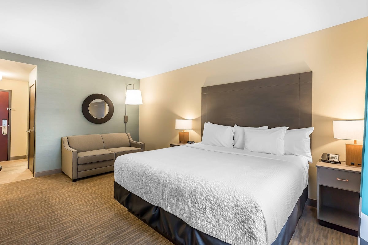 Une chambre d’hôtel avec un grand lit, un canapé, un miroir rond, deux tables de chevet avec des lampes et une porte donnant sur une autre pièce.