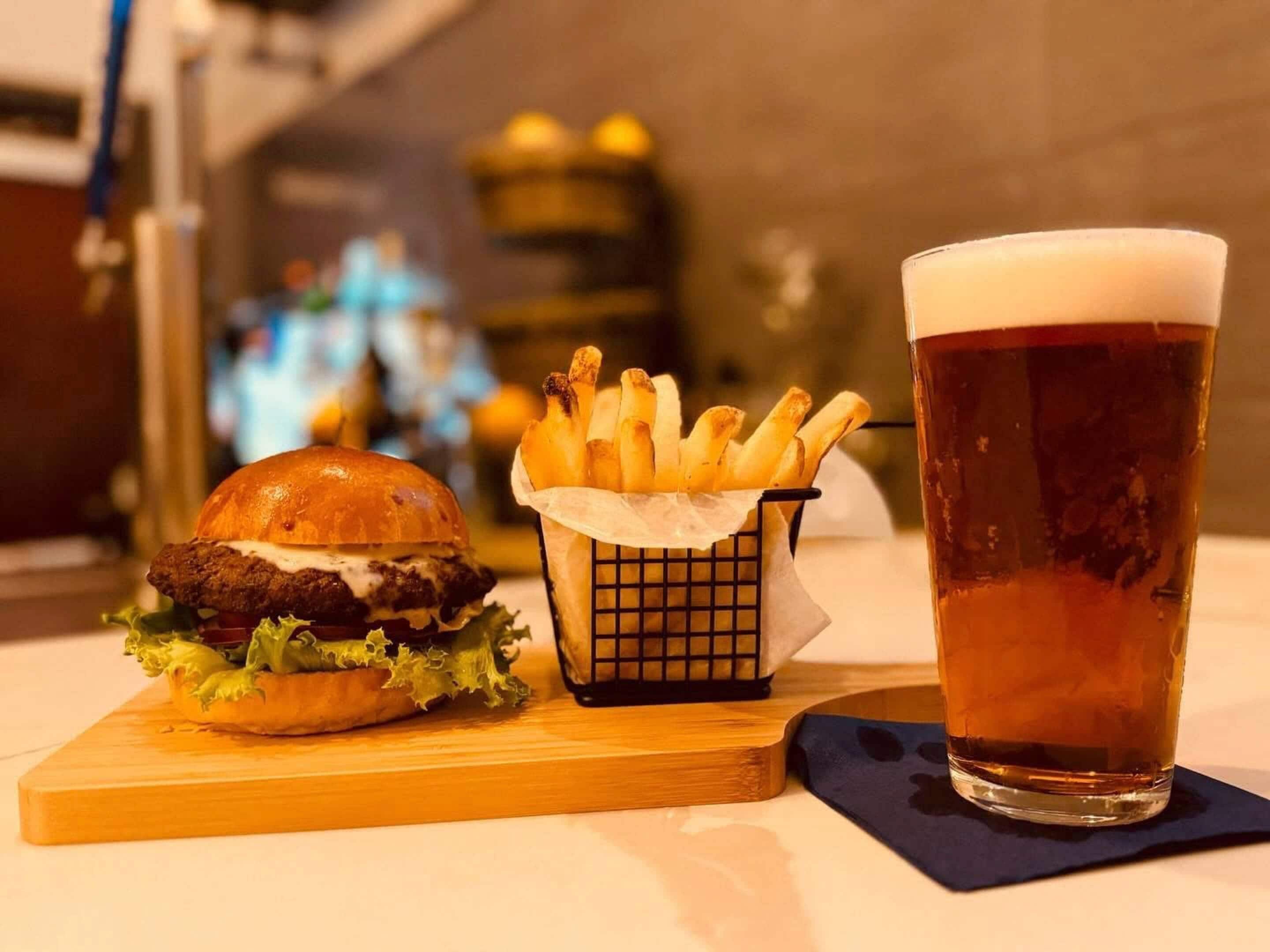 A burger, a basket of fries, and a glass of beer on a wooden board.