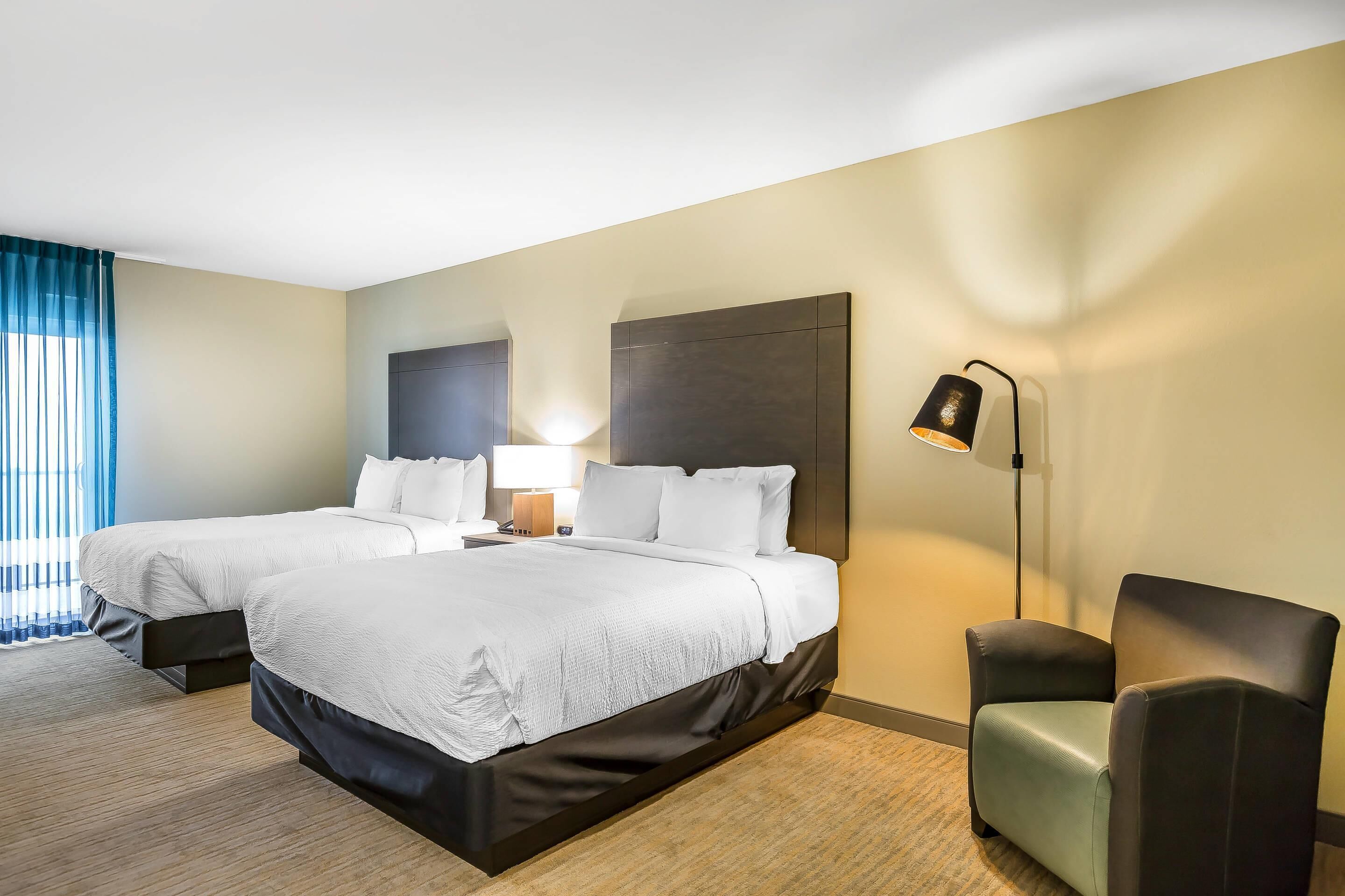 A hotel room with two beds, a desk, a chair, and a lamp.
