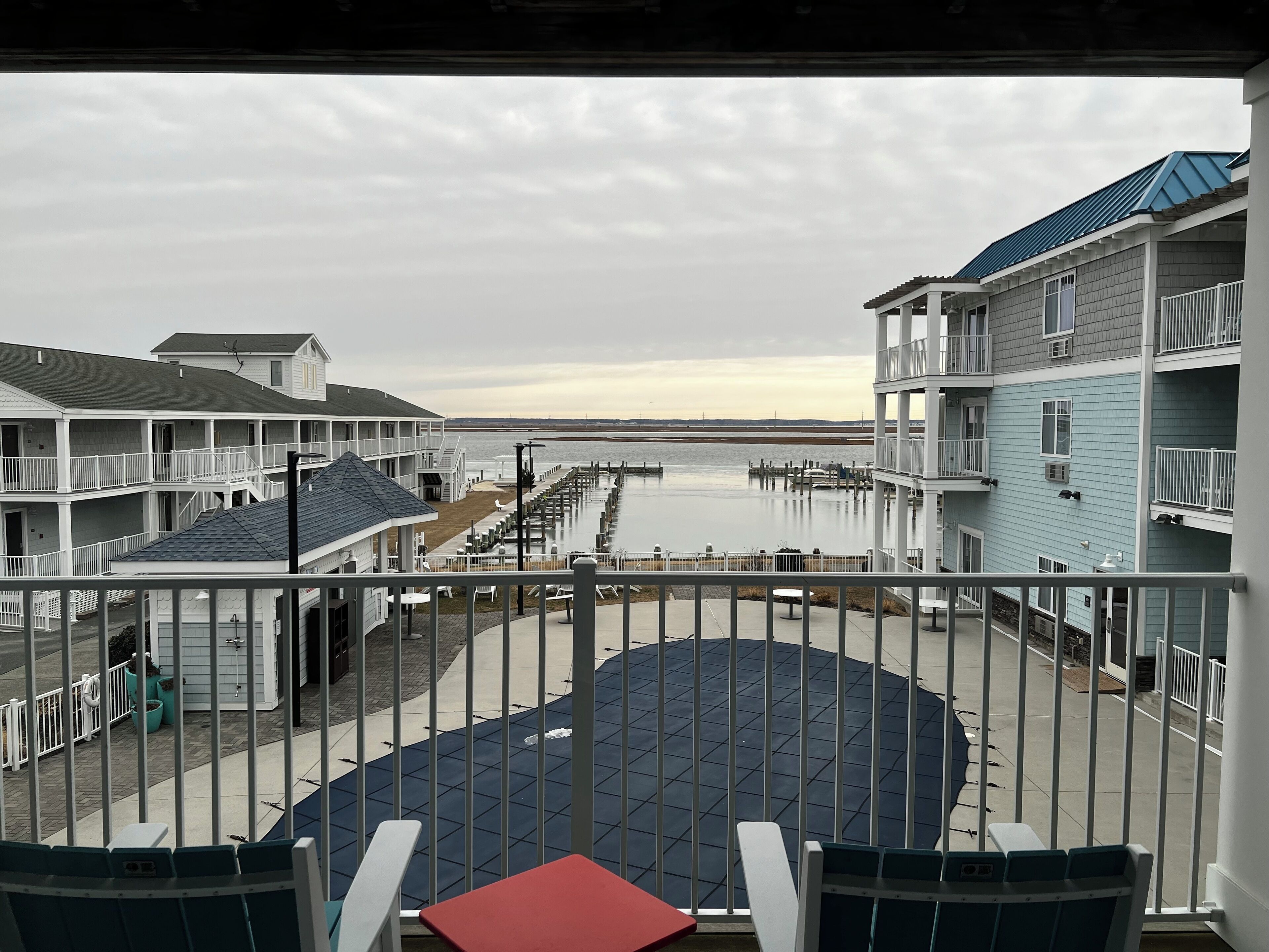 A view from a balcony overlooking a waterfront with a pier, featuring multi-story buildings with balconies and a red table in the foreground.