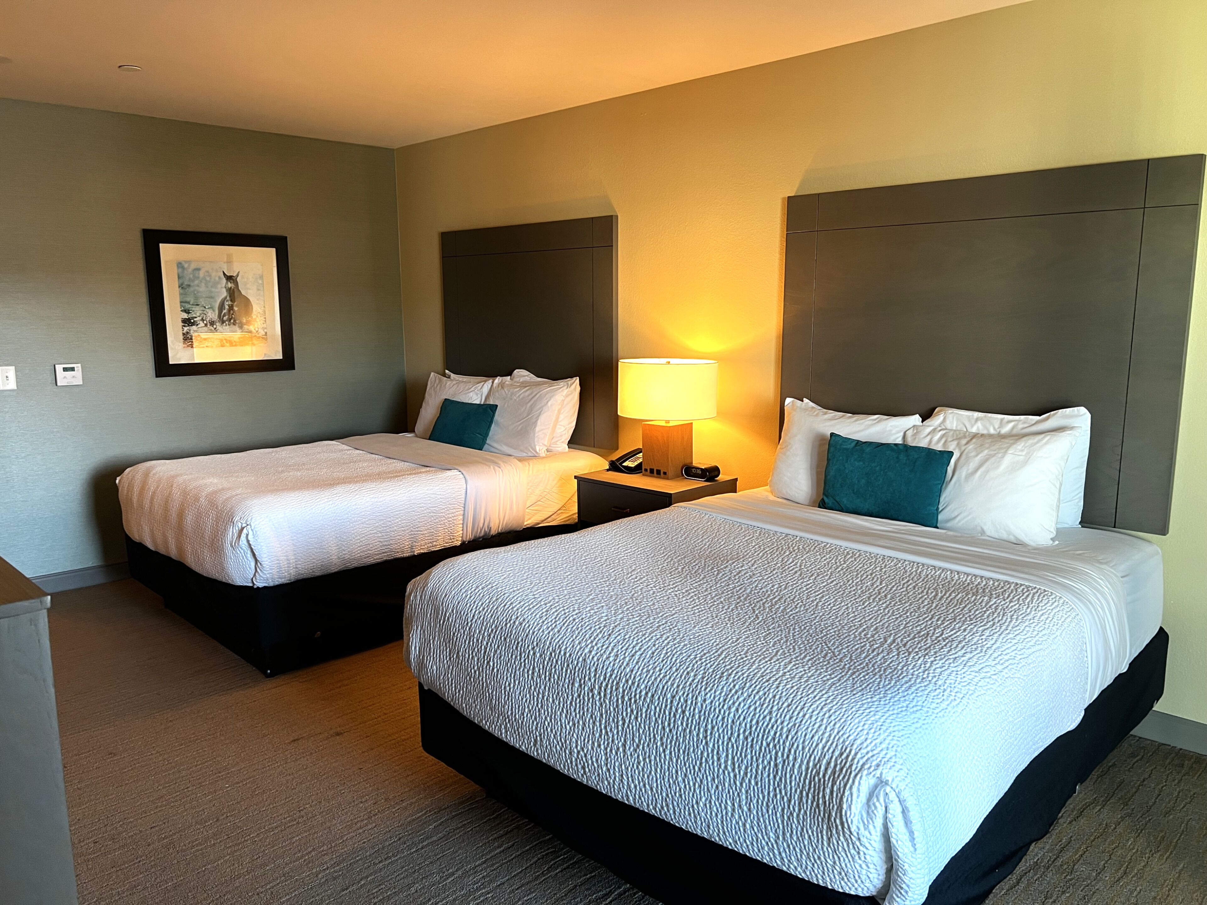A hotel room with two beds, a nightstand with a lamp, a framed picture on the wall, and a small desk.
