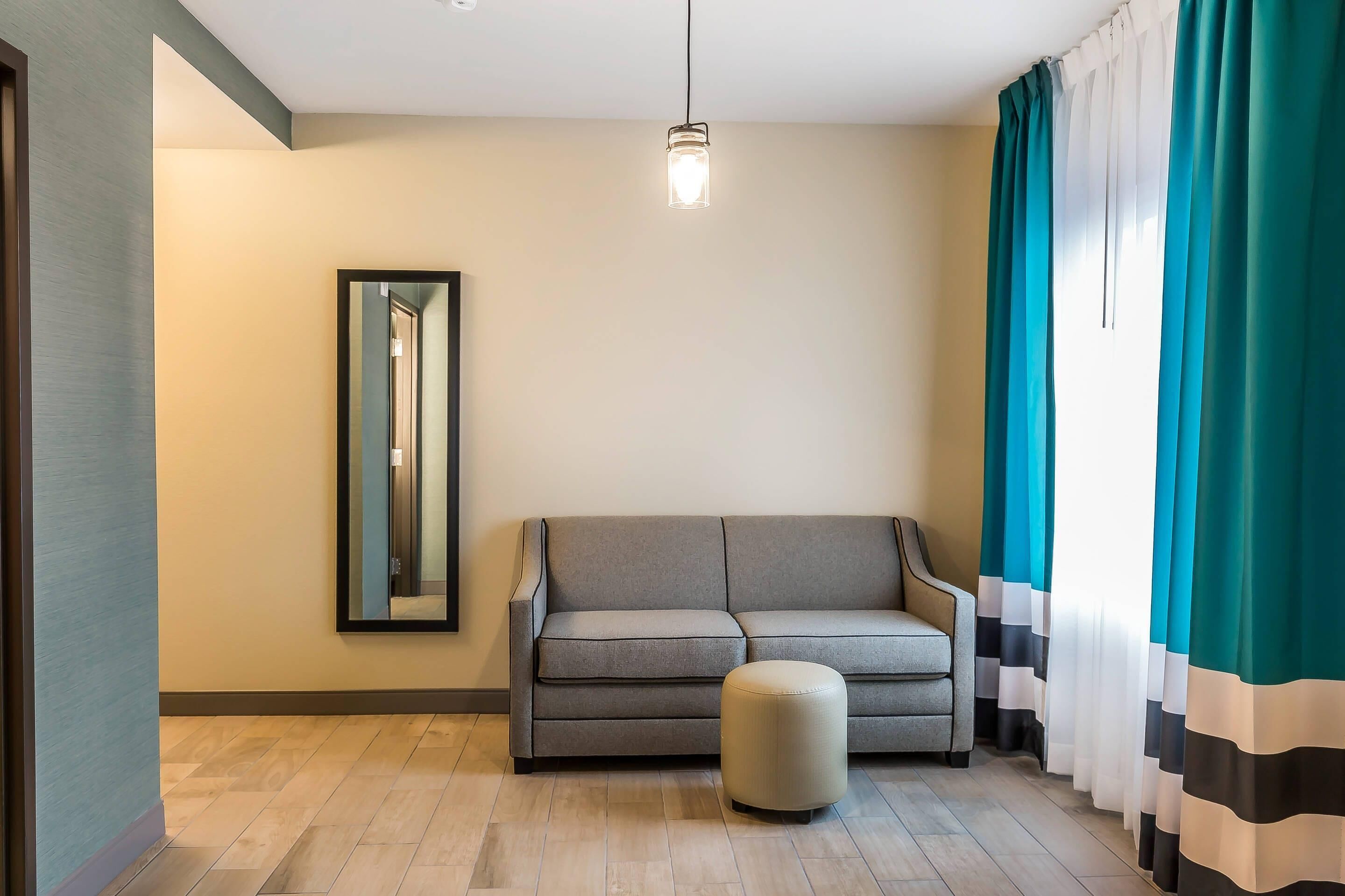 A modern living room with a grey sofa, a round ottoman, a large mirror on the wall, and teal curtains.