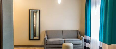 A modern living room with a grey sofa, a round ottoman, a large mirror on the wall, and teal curtains.