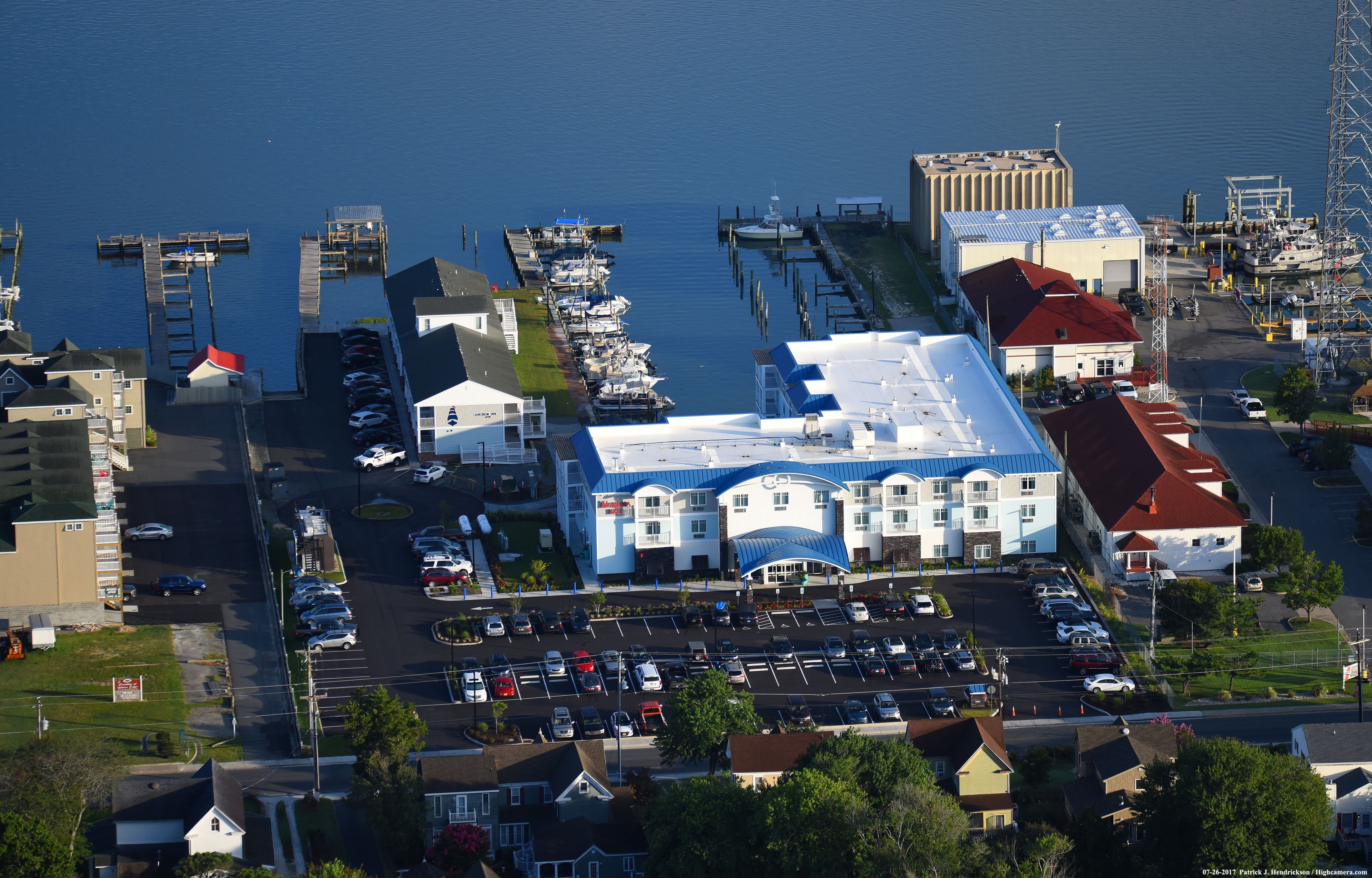 An aerial view of a marina with docked boats, a parking lot, and buildings with a red roof and blue awnings.