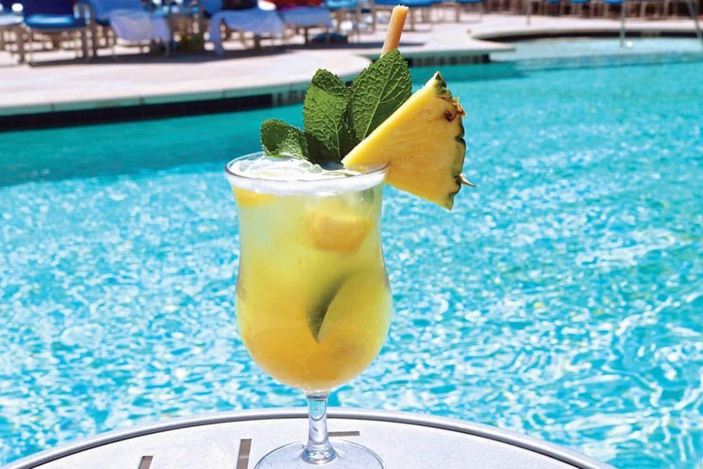 A cocktail with a pineapple garnish and mint leaves, placed on a table by a pool.