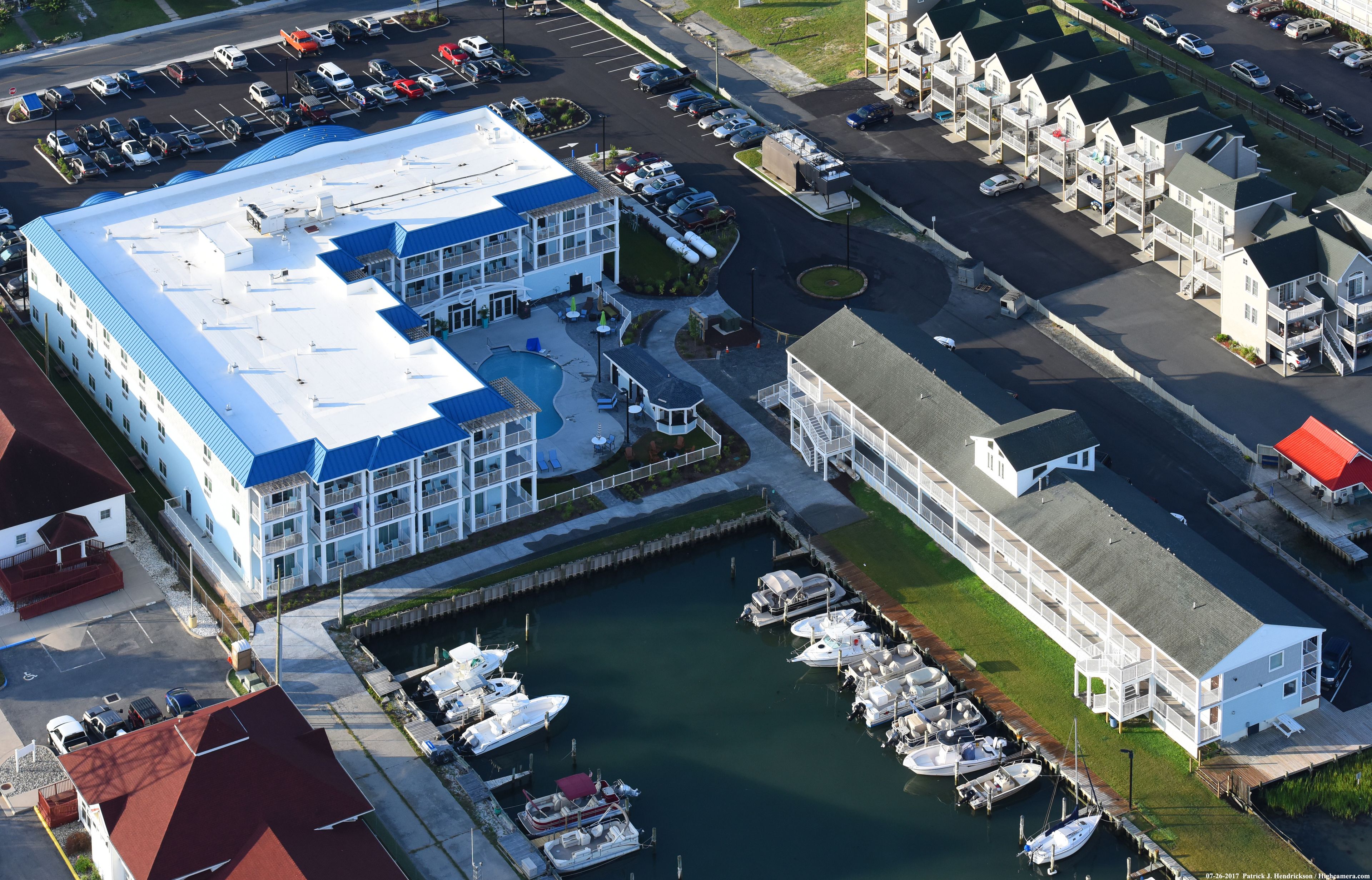 An aerial view of a marina with boats docked, a large building with a blue roof, and a parking lot with several cars.