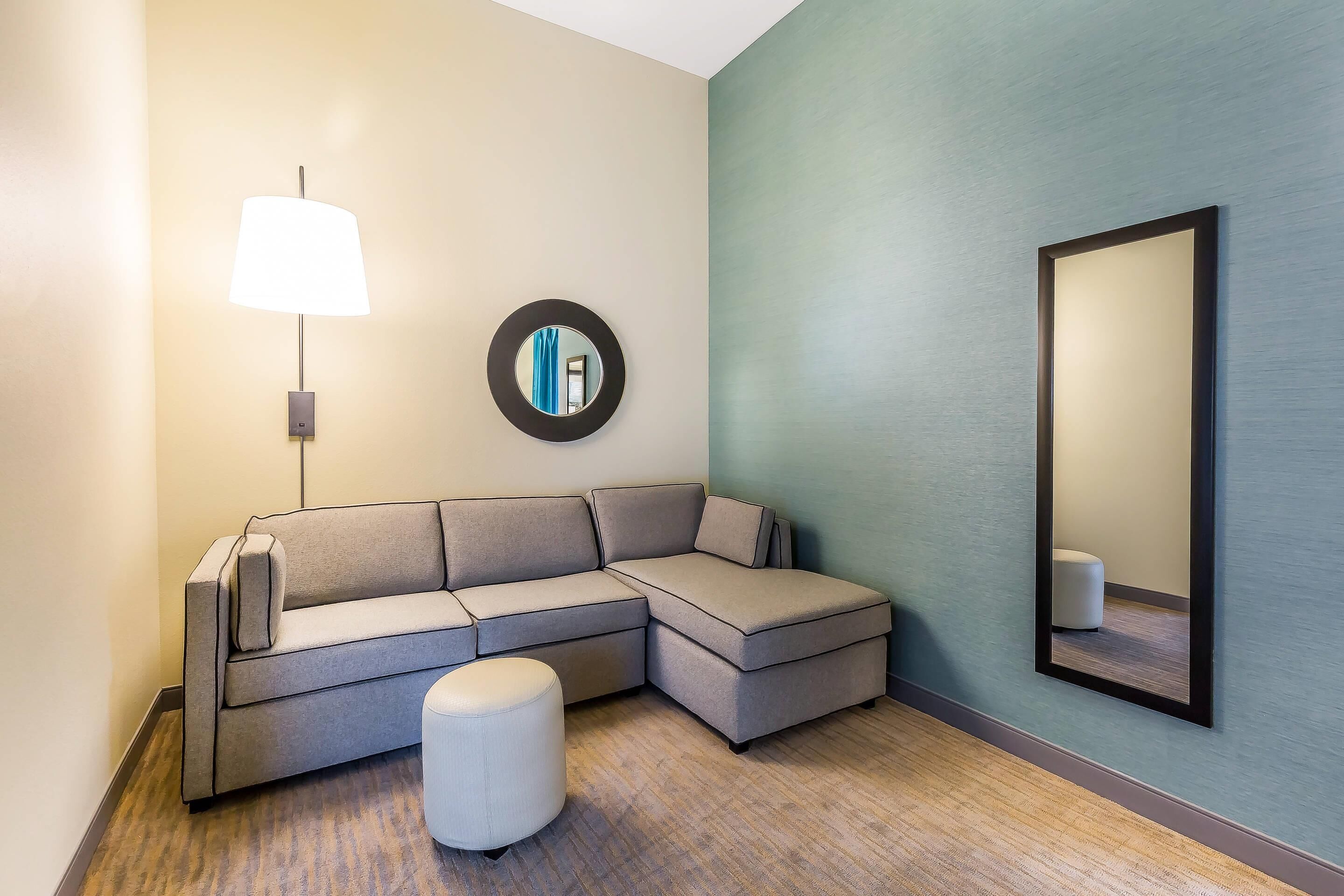 A modern hotel room with a sectional sofa, a round mirror, and a wall-mounted lamp.