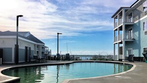 A pool area with a view of the water, a building with a balcony, and a clear sky.