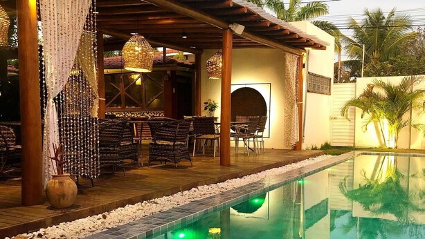 A poolside area with a tiled pool, wicker furniture, and a covered seating area with a hanging light fixture. - Pousada Le Baron (Marechal Deodoro)