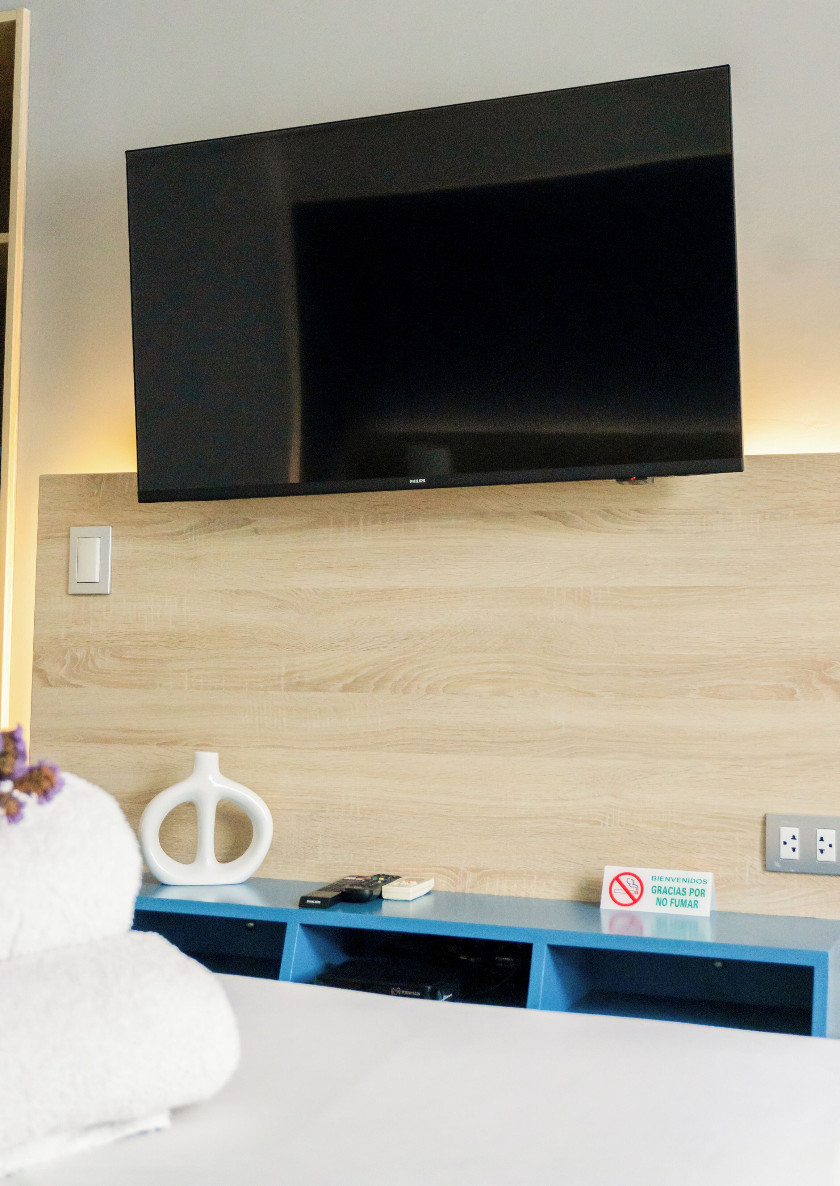 Habitación de hotel moderna con un televisor de pantalla plana montado en una pared de madera, un florero blanco, una pequeña mesa con un control remoto y un letrero que indica prohibido fumar.